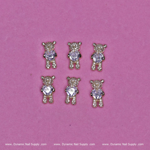 Six small teddy bear charms with a sparkling, gem-like appearance are set against a pink backdrop. Each charm features a clear body and head adorned with rhinestones, highlighted by gold detailing on the limbs and ears. The playful design beautifully embodies the style of Dynamic Nail Supply's (Gold) 6 pcs Bear Charm with Rhinestones (Hugging) Engraved, making these charms exceptionally eye-catching.