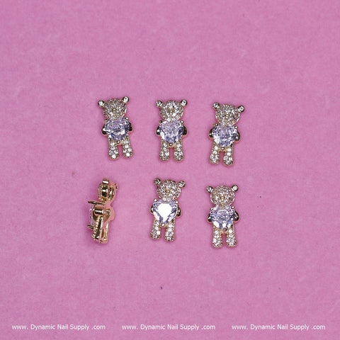Six small gold-toned bear charms with rhinestones, designed in a hugging pose, are displayed on a pink background. Five of the bears shine with their rhinestones, while one reveals its gold back. At the bottom, white text reads "www.DynamicNailSupply.com," showcasing where to purchase these delightful pieces from Dynamic Nail Supply.