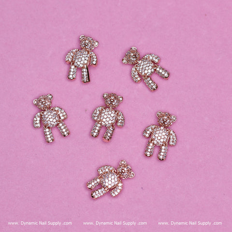 Six sparkling teddy bear charms, each adorned with tiny rhinestones, stand out against a pink background. These rose gold delights are perfect for those seeking unique nail art from Dynamic Nail Supply.