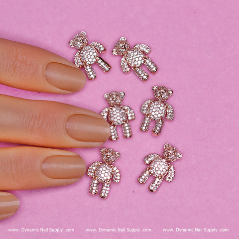 A hand with manicured nails painted in nude polish delicately places the (Rose Gold) 6 pcs Movable Teddy Bear Charm with Rhinestones Engraved on a pink surface. Each of the six identical charms sparkles with glistening rhinestones. The text Dynamic Nail Supply gracefully appears three times at the bottom edge.