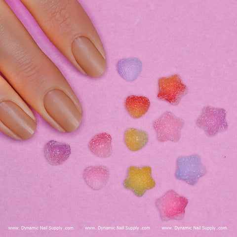 A manicured hand rests on a pink surface, decorated with vibrant, textured star and heart-shaped embellishments in shades of purple, pink, yellow, and red—ideal for any nail art design. The bottom of the image features "Dynamic Nail Supply" with the website www.DynamicNailSupply.com repeatedly in white text.