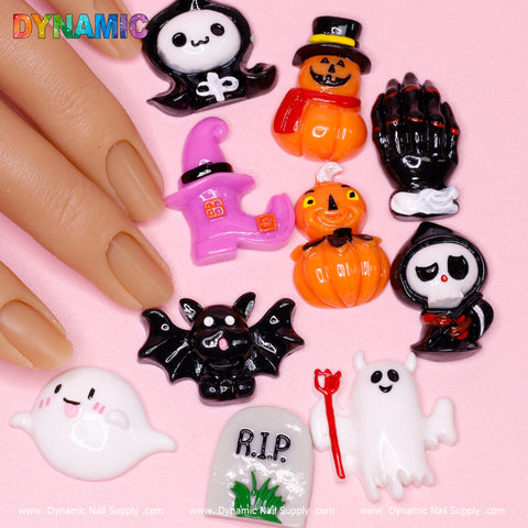 A collection of 10 adorable cartoon Halloween design nail charms featuring a ghost, bat, jack-o-lantern, skeleton, reaper, and more is set against a pink background. Beside the charms is a hand with manicured nails. The Dynamic Nail Supply logo is displayed in the top left corner.