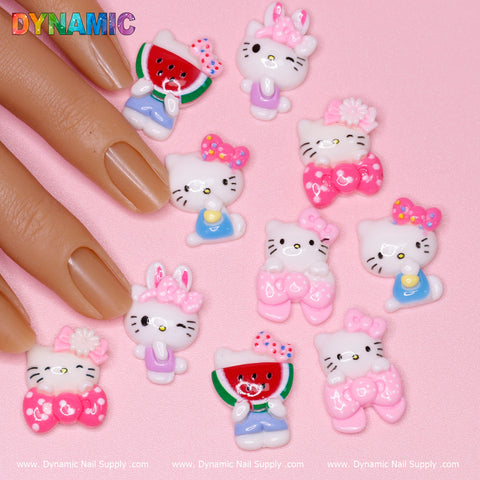 The 10 pcs Very Cute Hell0 Kitty Cat Family Design Nail Charms by Dynamic Nail Supply features cartoon-style cats in vibrant outfits and accessories such as bows and watermelons. Presented on a pink background with manicured nails, this whimsical display includes branding text at the bottom of the image.