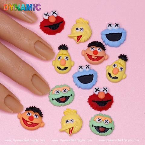 A hand with matte nude polish is shown near adorable 12 pcs Famous Sesame Street Cartoon Characters Nail Charms from Dynamic Nail Supply. These lively charms, featuring blue faces with wide mouths, yellow faces with fuzzy hair, and red or orange expressions, stand out against a pink background.