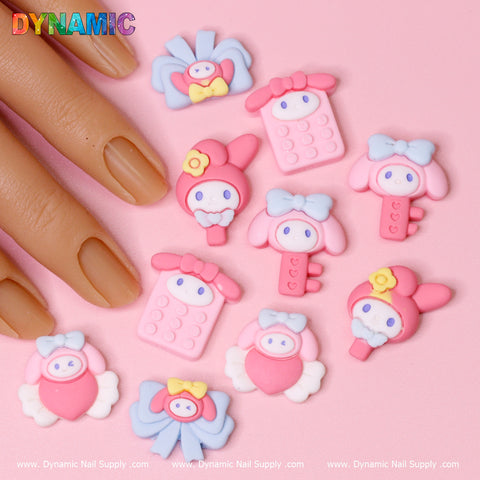 A set of pastel-colored 10 pcs Cute Hell0 Kitty Cat Design Nail Charms by Dynamic Nail Supply, displayed on a pink background. The adorable cartoon characters feature pink and blue bows and hold miniature phones. A hand with brown painted nails peeks in on the left side.
