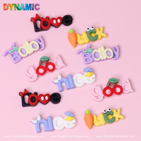 A vibrant assortment of clay-inspired, colorful words sits against a pink background, featuring the words Love, Baby, Duck, Nice, and Good adorned with kawaii cartoon nail charms such as hearts and bunnies. In the top left corner, the product brand name "Dynamic Nail Supply" is showcased in lively rainbow colors.