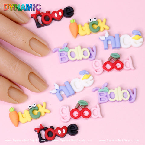 A vibrant display of 3D nail art decorations from Dynamic Nail Supply showcases the "10 pcs Cute Cartoon Letters Words Nail Charms" on a pink backdrop. The designs feature whimsical words like love and luck, as well as charming adornments like a carrot and starfish. A hand with neatly manicured nails is visible in the top left corner.