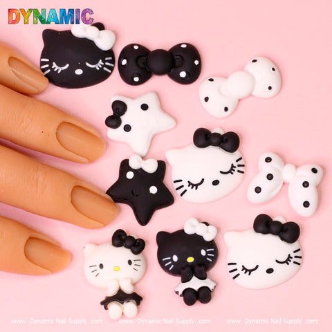 A hand with painted nails rests beside several adorable nail charms from the "10 pcs Cute Hello Kitty Cat Cartoon Design Nail Charms" collection by Dynamic Nail Supply on a pink surface. The charms showcase a playful assortment of black and white, cartoon-like designs featuring cats, bows, and stars, some accented with tiny faces. At the top left corner is an elegant script reading "Dynamic.