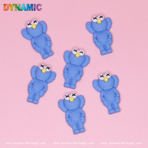Six blue KAWS Nail Designer Charms, featuring small cartoon characters with fluffy textures, crossed eyes, yellow noses, and outstretched arms, are arranged on a pink background. The word "DYNAMIC" is printed in colorful letters at the top left corner. Along the edge, you'll find "Dynamic Nail Supply" and "KAWS Nail Designer Charms" repeated like playful decorative elements.