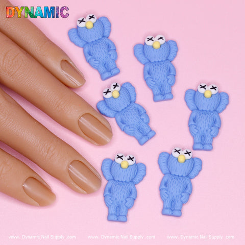 A close-up of a hand with smooth, neatly manicured nails in light nude rests on a pink surface, surrounded by six textured KAWS Nail Designer Charms (Blue) featuring crossed-out eyes and yellow noses. The top left showcases the word "DYNAMIC" from Dynamic Nail Supply.
