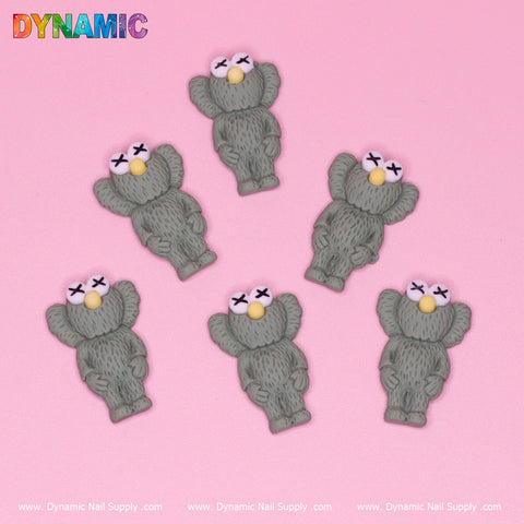 Six gray, cartoon-style bear figures with closed eyes and yellow noses are scattered on a pink background. The brand name, Dynamic Nail Supply, is displayed in colorful letters at the top left corner. Showcasing the 6 pcs KAWS Nail Designer Charms (Army Green), "Dynamic Nail Supply" text repeats along the bottom edge, making it perfect for nail art inspiration.