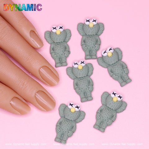 A hand with a natural manicure rests on a pink surface alongside six decorative pieces inspired by KAWS, styled as army green koalas with white eyes and textured fur. The top left showcases a vibrant Dynamic Nail Supply logo, adding flair to this charming display of nail art using the 6 pcs KAWS Nail Designer Charms.
