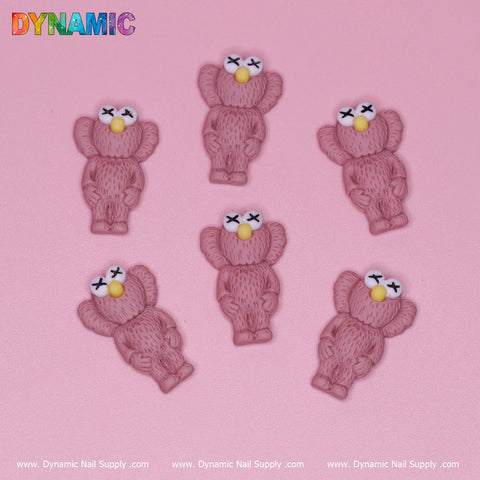 The image displays six KAWS Nail Designer Charms in the Pallet Slipper Pink color, shaped like koalas with yellow noses and X-shaped eyes, set against a pink backdrop. Each charm highlights intricate furry texture details. "DYNAMIC" is prominently placed in colorful letters at the top left corner, while www.DynamicNailSupply.com runs along the bottom edge.