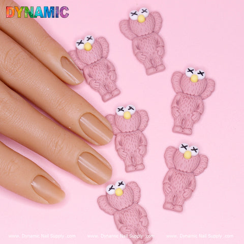 A hand with neatly manicured, natural-colored nails rests on a pink surface. Surrounding it are six pink, fluffy teddy bear nail charms featuring white eyes and yellow noses from the 6 pcs KAWS Nail Designer Charms (Pallet Slipper Pink) by Dynamic Nail Supply. The word DYNAMIC appears in colorful letters at the top left corner, with branding text at the bottom.