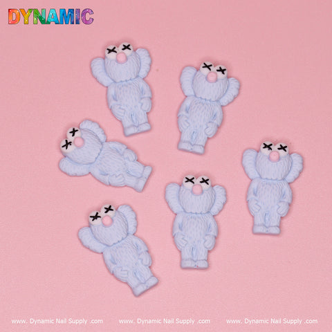 Six matching Baby Blue KAWS Nail Designer Charms are set in a circular formation against a pink backdrop, each featuring cartoon-like bear designs with crossed-out eyes and pink noses. The word "DYNAMIC" is displayed in vibrant letters at the top left, showcasing these playful charms from Dynamic Nail Supply along the bottom edge.