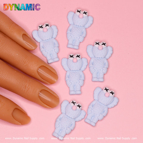 A close-up shot features a hand with light brown nails next to six bear-shaped nail charms in blue, crafted by Dynamic Nail Supply. These KAWS Nail Designer Charms have X-shaped eyes and round pink noses. The background is pink, adorned with the vibrant "DYNAMIC" text in the top left corner, showcasing the charm's creative design flair.