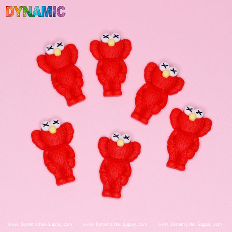 Six pieces of the 6 pcs KAWS Nail Designer Charms (Red) by Dynamic Nail Supply with crossed-out eyes and a cartoonish flair are artfully arranged on a pink background. These identical charms feature yellow noses and textured surfaces. The Dynamic Nail Supply logo shines brightly at the top, with the website conveniently displayed at the bottom.