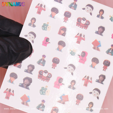 A person's hand in a black glove holds a sheet of small stickers from Dynamic Nail Supply against a pink background. These Squid Game-inspired stickers, titled "Squid game 01 (sq game)," feature colorful characters and objects such as people wearing square and circle masks, geometric shapes, and cartoonish faces, all arranged in neat rows.
