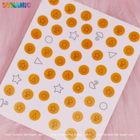A sheet of nail art stickers features orange circles adorned with black question marks, capturing the enigmatic essence of "Squid Game." Black outlined shapes such as triangles, stars, and crescent moons add an element of intrigue against a gentle pink background. The Dynamic Nail Supply brand name is prominently displayed at the bottom.