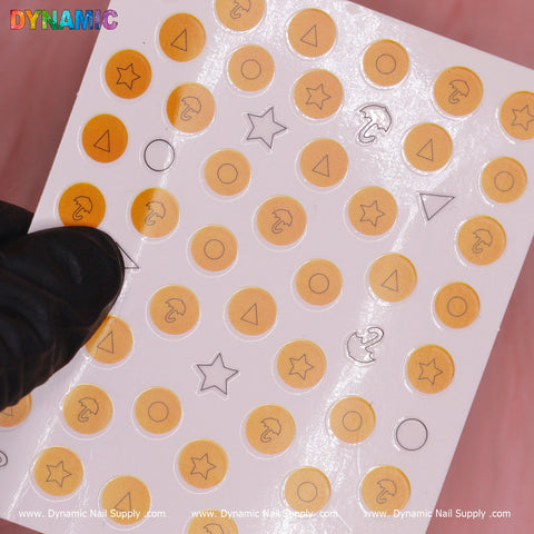 A sheet of nail decals is displayed, showcasing designs such as circles, triangles, stars, and semicircles in yellow with black outlines, evoking the style from Squid Game. A hand wearing a black glove holds the sheet against a pink background. The bottom features repeated text: Dynamic Nail Supply. The product is named "Squid game 02 (sq game)" by Dynamic Nail Supply.