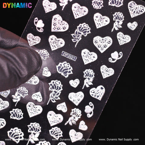 A gloved hand holds a transparent sheet showcasing white nail decals featuring hearts, flowers, and roses. The product is labeled as heart, flower, rose (white) (DP 2021), with the background displaying the URL www.DynamicNailSupply.com and the brand name Dynamic Nail Supply in vibrant text.