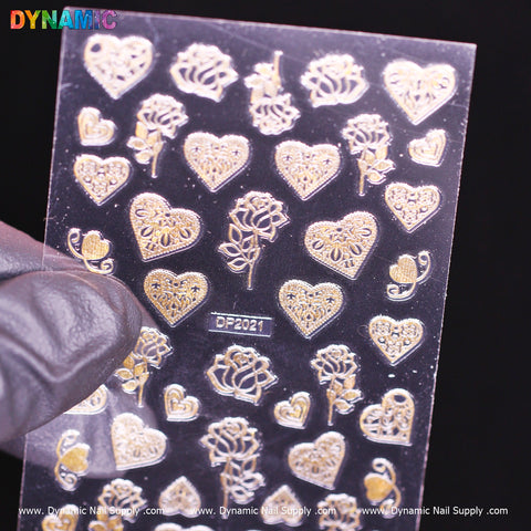 A gloved hand holds a transparent sheet featuring Dynamic Nail Supply's intricate rose gold nail art stickers from the "heart, flower, rose (gold) (DP 2021)" collection. The designs include heart shapes and ornate lace-like patterns, elegantly standing out against the black background with the product code DP2021 subtly printed on the sheet.