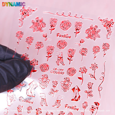 A hand wearing a black glove holds a hand writing art (red) nail art sticker sheet (CB 199) from Dynamic Nail Supply, featuring transparent red designs. The sheet showcases patterns such as roses, high heels, and the word "FASHION" in decorative fonts. Set against a soft pink background with Dynamic Nail Supply branding, this product truly represents handwriting art for your nails.