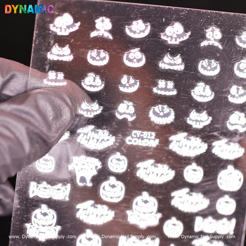 A gloved hand holds a sheet of Halloween glow-in-the-dark nail decals from Dynamic Nail Supply. The sheet features spooky designs such as skulls, pumpkins, ghosts, and bats. The word DYNAMIC is visible at the top in colorful text with CY 012, while www.DynamicNailSupply.com is printed at the bottom.
