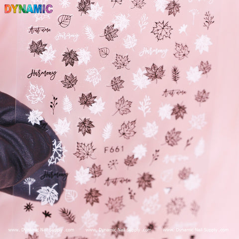A gloved hand elegantly presents a sheet of nail art stickers from Dynamic Nail Supply, featuring the "fall leaves (black and white) (F 661)" design collection against a soft pink backdrop, beautifully capturing the essence of seasonal beauty with harmonious autumn-themed patterns.