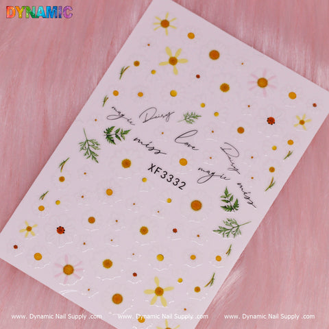 On a pink surface rests a white nail art sticker sheet labeled as White Daisy Flower (XF 3332) from Dynamic Nail Supply, featuring various designs such as yellow daisies, white daisy flowers with orange centers, green leaves, and cursive words like "love" and "miss you.