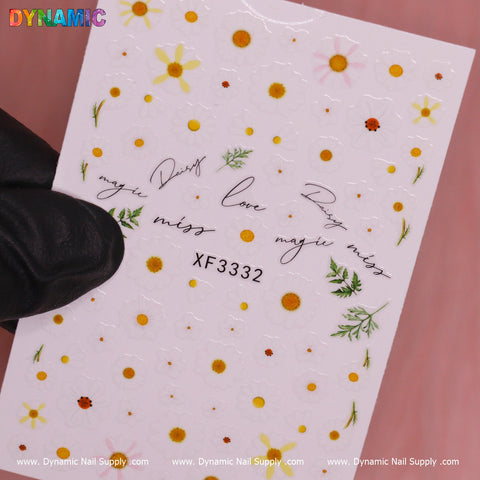 A sheet of nail decals from Dynamic Nail Supply, named White Daisy Flower (XF 3332), showcases glossy designs of small white daisies with scattered green leaves. Black cursive words such as love, miss, and dream are interspersed amongst the flowers. The sheet is held by a gloved hand, prominently displaying the model number XF 3332 in the center.