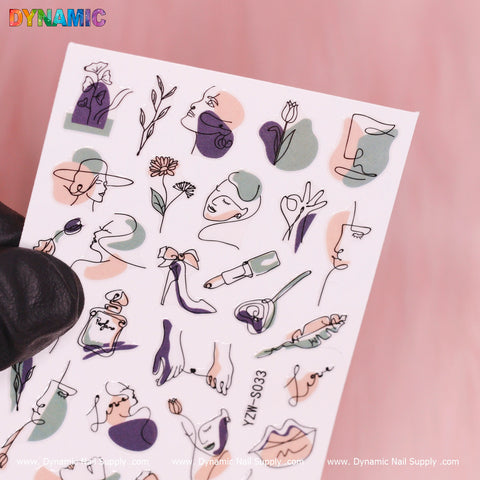A gloved hand holds a sheet of Dynamic Nail Supply's Hand Writing (YZW S033) nail art stickers, showcasing abstract designs such as faces, flowers, and perfume bottles. The shades of pink, green, and black beautifully complement the soft pink background.