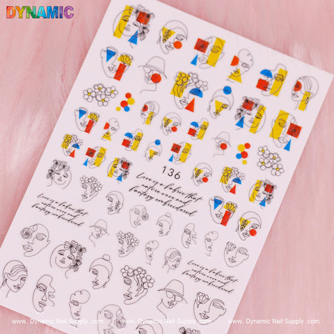 A sheet of "Hand Writing Face (136)" nail art stickers from Dynamic Nail Supply is displayed against a pink background. The designs feature a mix of colorful abstract faces with geometric shapes, flowers, and hats, alongside monochrome outlines. In the top left corner, the word "DYNAMIC" appears in vibrant letters, with the number 136 and artistic instructions centrally written in an elegant script.