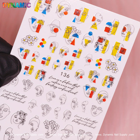 A close-up of the Hand Writing Face (136) sheet from Dynamic Nail Supply features decorative nail stickers with abstract line art faces, a touch of handwriting elegance, floral patterns, and whimsical drink illustrations. Some stickers are embellished in red, yellow, and blue hues. The number 136 is prominently displayed against a soft pink background.