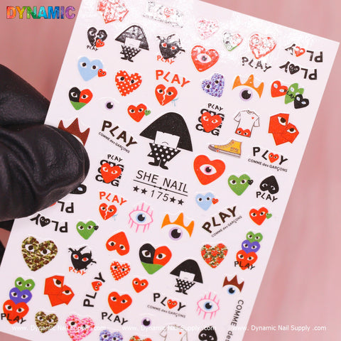 This lively sticker sheet from Dynamic Nail Supply, the Comme Des Garcons Sticker (175), showcases colorful nail decals with whimsical designs such as hearts, eyes, strawberries, and bears. Personalize your nails with text like COMME des GARÇONS and PLAY. Some hearts are embellished with curious eyes while polka dots and stripes enhance the overall charm. A gloved hand displays the sheet against a pink backdrop.