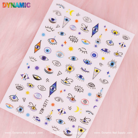 The Evil Eyes and Spirit styles nail art stickers from Dynamic Nail Supply (Model: YZW S271) feature vibrant designs that include stars, crescent moons, sunburst patterns, and both open and closed eyes with lashes. Available in eye-catching shades of blue, purple, yellow, and pink with the word "DYNAMIC" in rainbow colors adorning the top left corner.