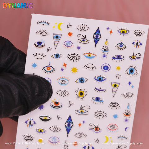 A hand in a black glove holds a sheet of Evil Eyes and Spirit styles (YZW S271) nail stickers, which features various eye designs, moons, stars, and geometric shapes. In the top left corner, the word "DYNAMIC" appears in vibrant letters. The background has a soft pink hue that captivates one's attention with its spirit styles.