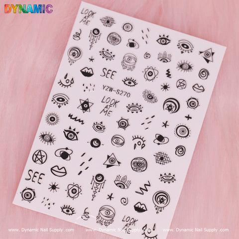 A sheet of Evil Eyes and Spirit styles (Black and White) (YZW S270) nail art stickers from Dynamic Nail Supply is set against a pink background, highlighting a variety of black designs such as eyes, stars, moons, circles, and abstract shapes. Some stickers incorporate Spirit styles with phrases like "LOOK AT ME" and "SEE," with the brand's logo visible along the edges of the sheet.