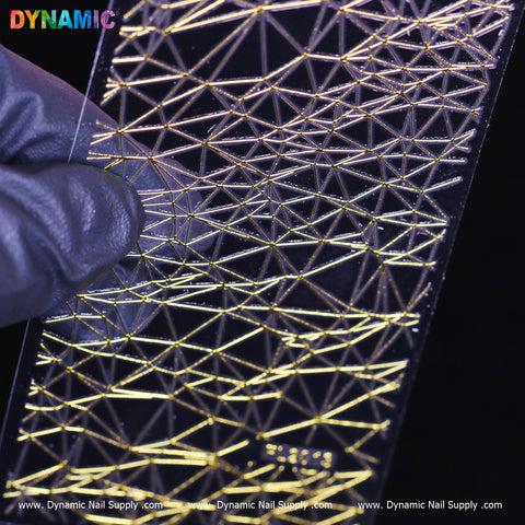 A gloved hand holds a transparent nail stencil by Dynamic Nail Supply, showcasing the Gold Net Texture Geometric (FL 2013) design with intricate patterns of triangles and lines. The dark background emphasizes the reflective quality of this stylish and modern aesthetic, perfect for nail art.