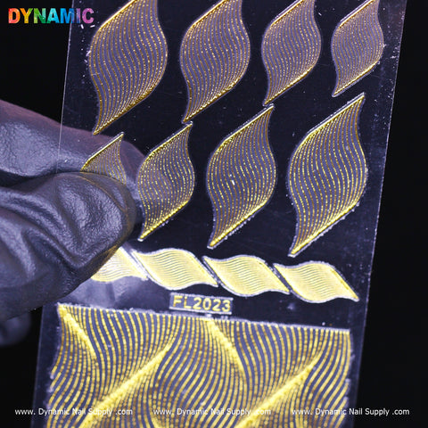 A gloved hand holds a transparent sheet featuring gold nail art stickers shaped like abstract leaves, intricately designed with gold swirl patterns. The sheet is labeled as Gold swirl pattern (FL 2023) and prominently displays the Dynamic Nail Supply branding multiple times.