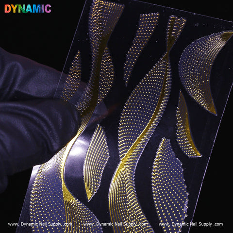 A gloved hand holds a transparent sheet showcasing intricate gold nail art decals adorned with elegant swirl patterns and gold dots. Set against a dark background that enhances the shimmer of the details, the brand name, Dynamic Nail Supply, appears at both the top and bottom of the sheet in a style reminiscent of their Gold dot and swirl pattern (FL 2020) design.