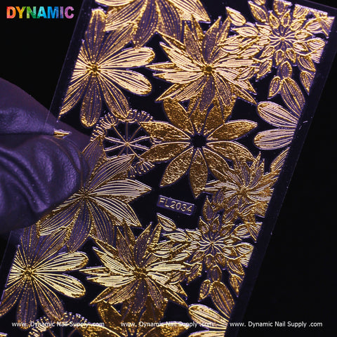 A hand holds a shiny rectangular sheet featuring intricate gold floral designs against a dark background. The top left corner proudly displays "Dynamic Nail Supply" in vibrant letters, and at the bottom, the elegant presentation of the Gold Flowers (FL 2034) website URLs can be seen.