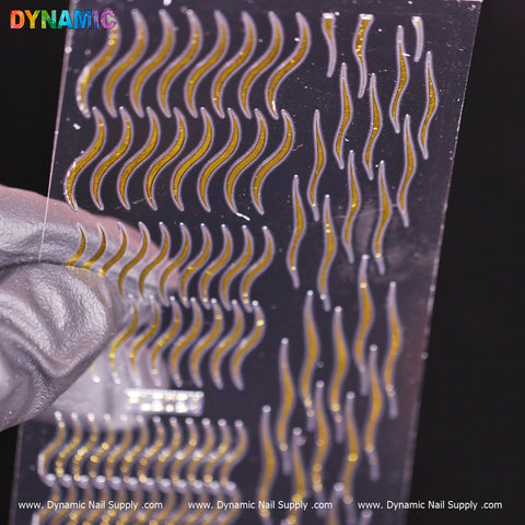 A gloved hand holds a transparent sheet featuring the Gold parallel line wave pattern nail stickers in various sizes. The product, from Dynamic Nail Supply, gleams against a dark background, highlighting its shiny texture. At the bottom, the company's website URL and "FL 2024" are visible, indicating its latest collection.