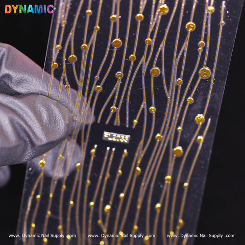 A gloved hand holds a clear plastic sheet featuring intricate gold decals with thin, wavy lines and small dots, showcasing the Gold Abstract design pattern by Dynamic Nail Supply. Against a dark background that highlights the shiny gold details, the colorful "Dynamic FL 2033" text is visible at the top left.