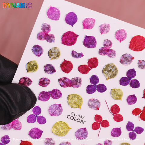 A close-up shot showcases a hand in a black glove holding a sheet of vibrant nail art stickers featuring small leaf shapes. The stickers are designed with shades of pink, red, and purple leaves accented by hints of gold. The text at the bottom reads "CL 037" from the Purple Pink Red Leaves collection by Dynamic Nail Supply, bordered by the brand's logos along the sheets' edges.