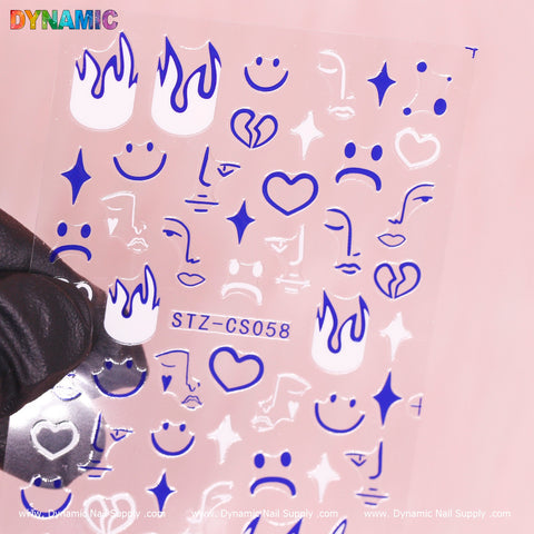A gloved hand holds a transparent sheet of nail art decals featuring the Hand writing Abstract Face Emoji (STZ CS058) by Dynamic Nail Supply. The sheet showcases blue icons, including flames, abstract face emojis, hearts, stars, and musical notes. In the top left corner, "Dynamic" is boldly written in colorful letters above the designs.