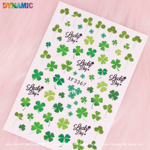 A sheet of Clovers and Lucky Leaves (XF 3367) nail stickers is displayed on a light pink background. The green clover designs come in various sizes and shapes, with some labeled Lucky Day. The branding at the bottom reads Dynamic Nail Supply. It's an ideal addition to your Lucky Leaves collection!