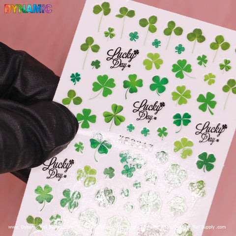 In a gloved hand, a sheet of Dynamic Nail Supply's Clovers and Lucky Leaves (XF 3367) nail decals is displayed, featuring an array of shamrocks and clovers alongside the phrase "Lucky Day" in elegant black script. The clovers come in various sizes and styles, with some presenting solid green colors while others gleam with a reflective metallic sheen against the light pink backdrop.