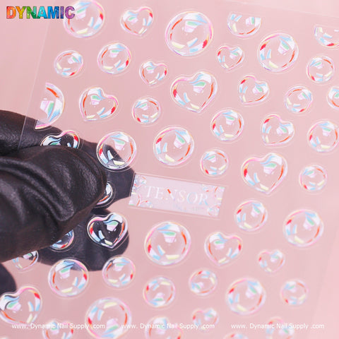 A gloved hand is holding a transparent sheet adorned with 3D Bubbles Iridescent stickers (TS 463) by Dynamic Nail Supply. The heart-shaped stickers vary in size, showing off a rainbow-like reflection against the soft pink background.