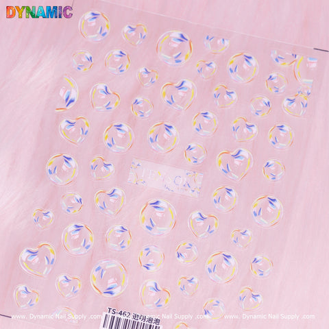 A sticker sheet featuring heart and circular holographic stickers is set against a pink, fluffy background. The iridescent surface shimmers with rainbow-colored hues, creating an enchanting effect. The phrase "Dynamic" adds a playful touch in colorful letters at the top left corner, highlighting the brand Dynamic Nail Supply's product "3D Bubbles Iridescent (TS 462).